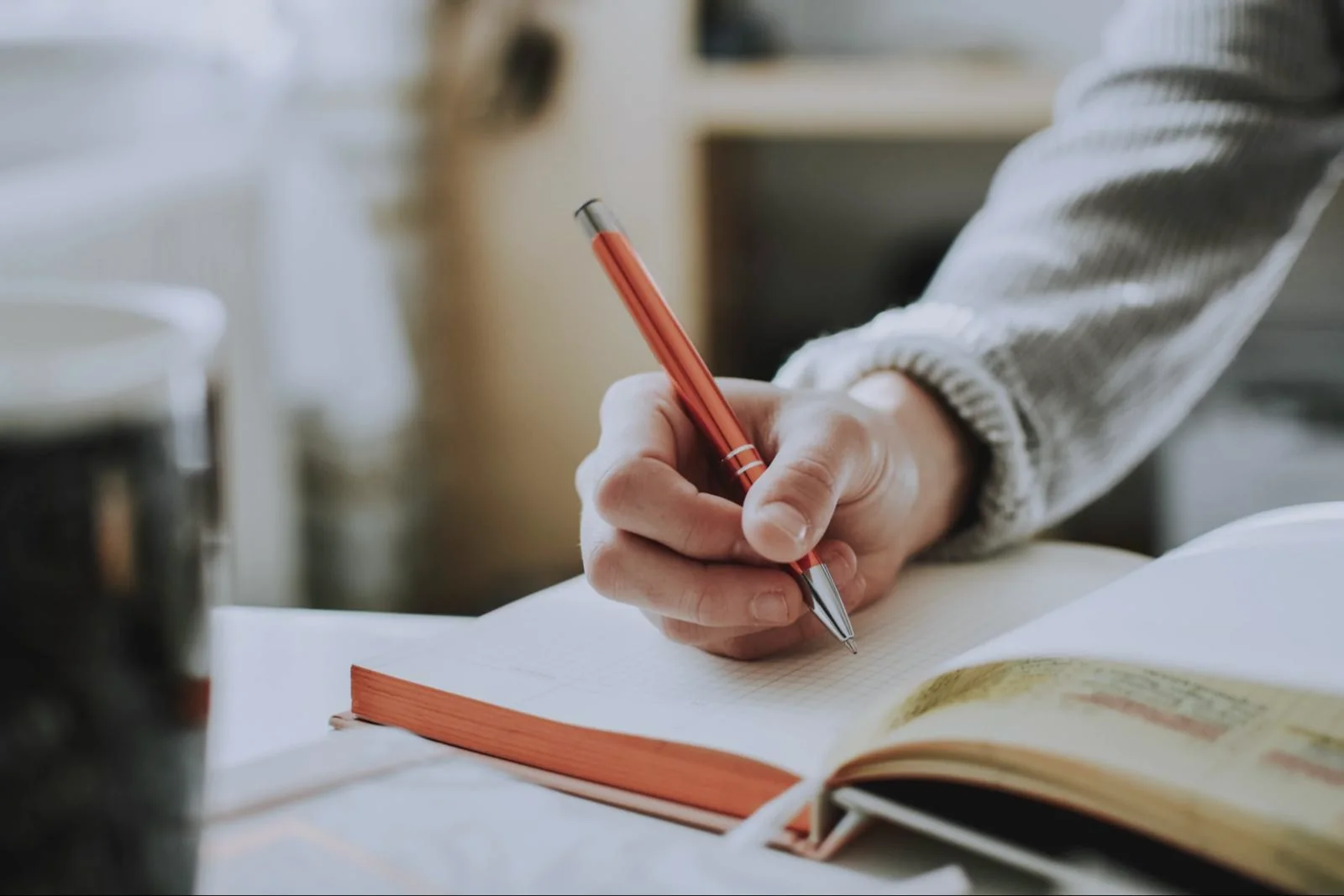 A person in a grey sweater writing in a notebook at a table in soft daylight.